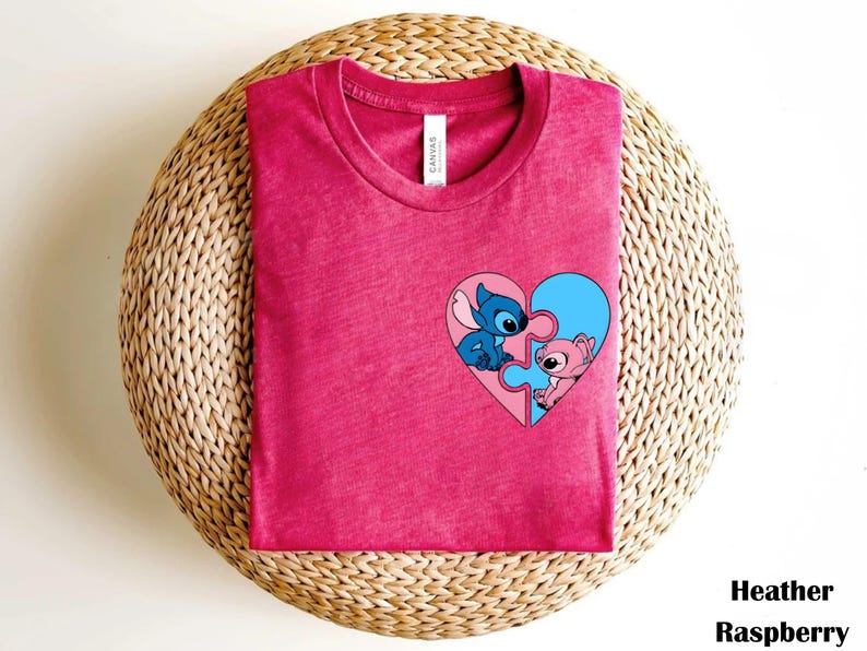 A pink t-shirt with a heart-shaped graphic featuring two cartoon characters, one blue and one pink, is displayed on a woven basket.