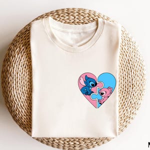 A white t-shirt with a heart-shaped graphic featuring two cartoon characters, one blue and one pink, on a woven basket.