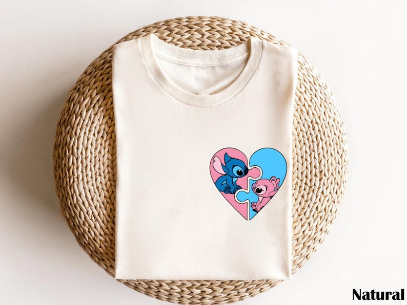 A white t-shirt with a heart-shaped graphic featuring two cartoon characters, one blue and one pink, on a woven basket.
