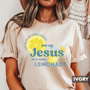 May include: Ivory-colored t-shirt with the text "Life gave me some lemons but my Jesus He be makin' LEMONADE" and a graphic of a lemon and a glass of lemonade. The shirt has rolled-up sleeves.