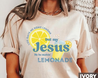 May include: Ivory-colored t-shirt with the text "Life gave me some lemons but my Jesus He be makin' LEMONADE" and a graphic of a lemon and a glass of lemonade. The shirt has rolled-up sleeves.