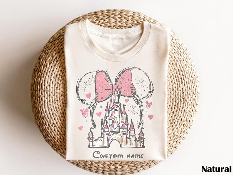 A white t-shirt with a pink bow and a castle illustration printed on it, placed on a woven basket.
