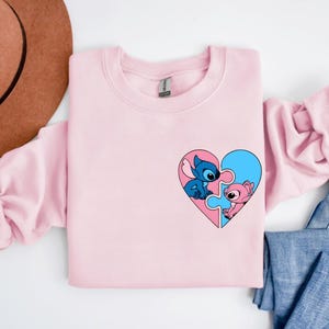 A pink long-sleeved shirt with a heart-shaped graphic featuring two cartoon characters, one blue and one pink, is displayed on a white background.
