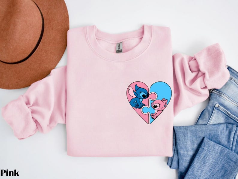 A pink long-sleeved shirt with a heart-shaped graphic featuring two cartoon characters, one blue and one pink, is displayed on a white background.