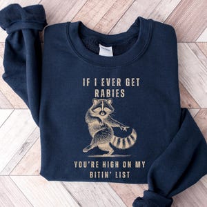 A navy blue sweatshirt with a graphic of a raccoon and text that reads "IF I EVER GET RABIES YOU'RE HIGH ON MY BITIN' LIST".