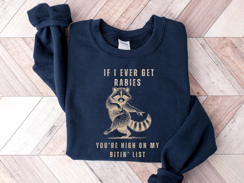 A navy blue sweatshirt with a graphic of a raccoon and text that reads "IF I EVER GET RABIES YOU'RE HIGH ON MY BITIN' LIST".