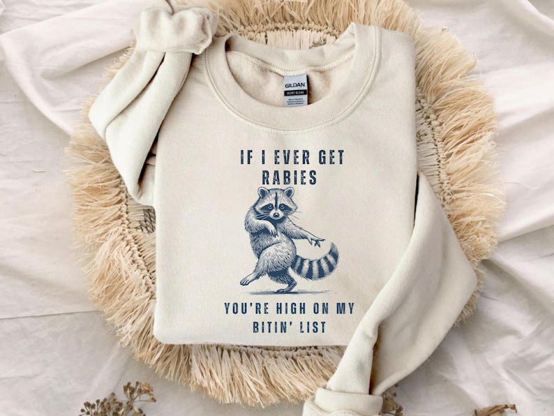 a white sweatshirt with a graphic of a raccoon and text that reads "If I ever get Rabies, you're high on my bitin' list".