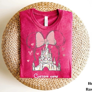 A pink t-shirt with a castle graphic and a pink bow on it, placed on a woven basket.