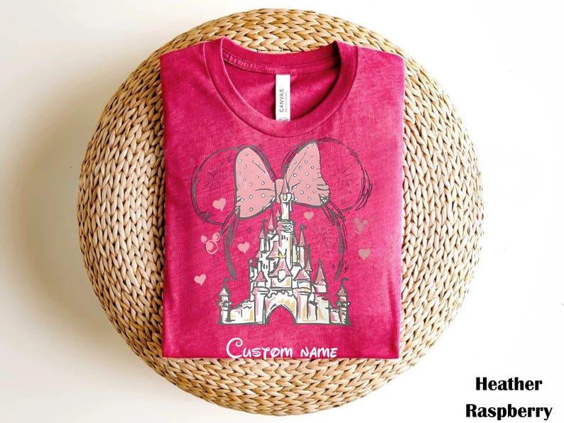 A pink t-shirt with a castle graphic and a pink bow on it, placed on a woven basket.