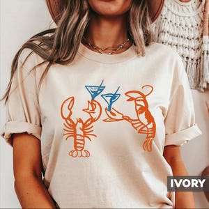 May include: Ivory-colored t-shirt featuring an orange lobster design holding blue cocktail glasses. The design is a playful illustration, perfect for casual wear. The word "IVORY" is printed in a white box at the bottom right.