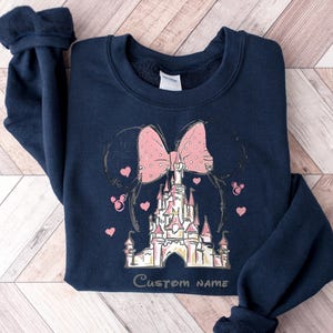 A navy blue sweatshirt with a pink bow and a castle graphic printed on it, along with the text "Custom Name" printed below the castle.
