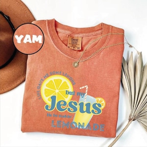May include: Coral-colored t-shirt with the text "Life gave me some lemons but my Jesus He be makin' Lemonade" and a graphic of a lemon and lemonade. A gold necklace and a brown hat are also visible. The shirt is a Comfort Colors brand.