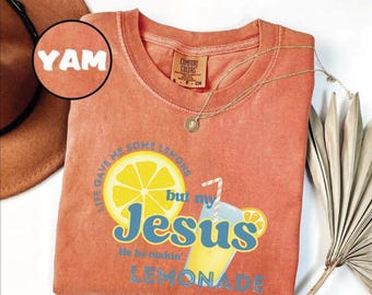 May include: Coral-colored t-shirt with the text "Life gave me some lemons but my Jesus He be makin' Lemonade" and a graphic of a lemon and lemonade. A gold necklace and a brown hat are also visible. The shirt is a Comfort Colors brand.