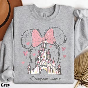 A gray sweatshirt with a pink bow and a castle graphic, along with a brown hat and a fan, are displayed on a white background.