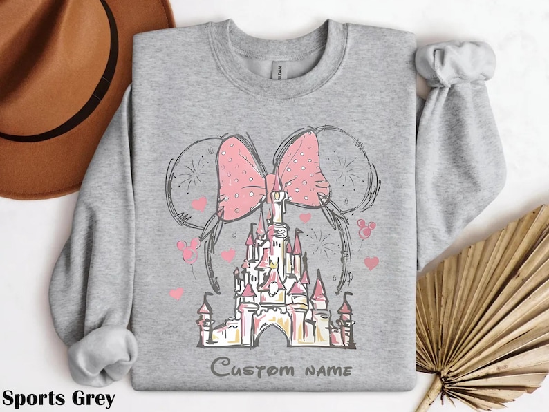 A gray sweatshirt with a pink bow and a castle graphic, along with a brown hat and a fan, are displayed on a white background.