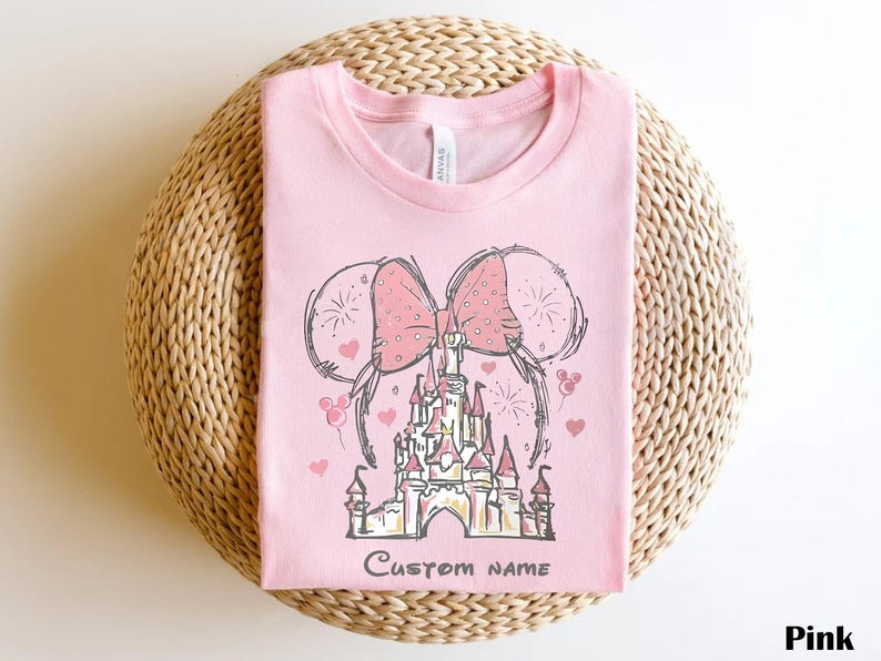 A pink t-shirt with a drawing of a castle and two pink bows, along with the text "Custom Name" printed on it.