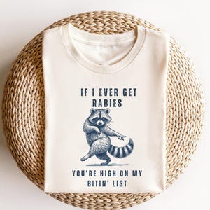 A white t-shirt with a graphic design of a raccoon and text that reads "IF I EVER GET RABIES YOU'RE HIGHLY ON MY BITIN' LIST".