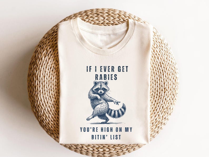 A white t-shirt with a graphic design of a raccoon and text that reads "IF I EVER GET RABIES YOU'RE HIGHLY ON MY BITIN' LIST".