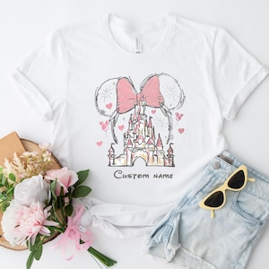 A white t-shirt with a pink bow and a castle graphic, along with a bouquet of pink flowers, a notebook, and a pair of sunglasses, all arranged on a white background.