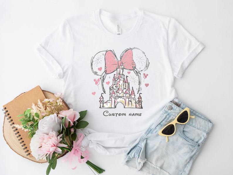 A white t-shirt with a pink bow and a castle graphic, along with a bouquet of pink flowers, a notebook, and a pair of sunglasses, all arranged on a white background.