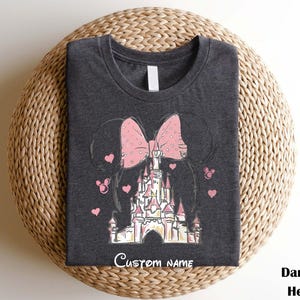 A gray t-shirt with a Disney castle design and a pink bow on it, placed on a woven basket.