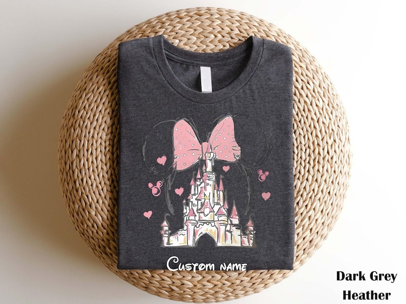 A gray t-shirt with a Disney castle design and a pink bow on it, placed on a woven basket.