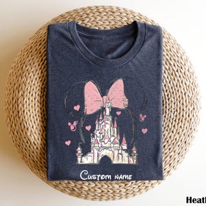 A dark blue t-shirt with a pink bow and a castle graphic printed on it, placed on a woven basket.