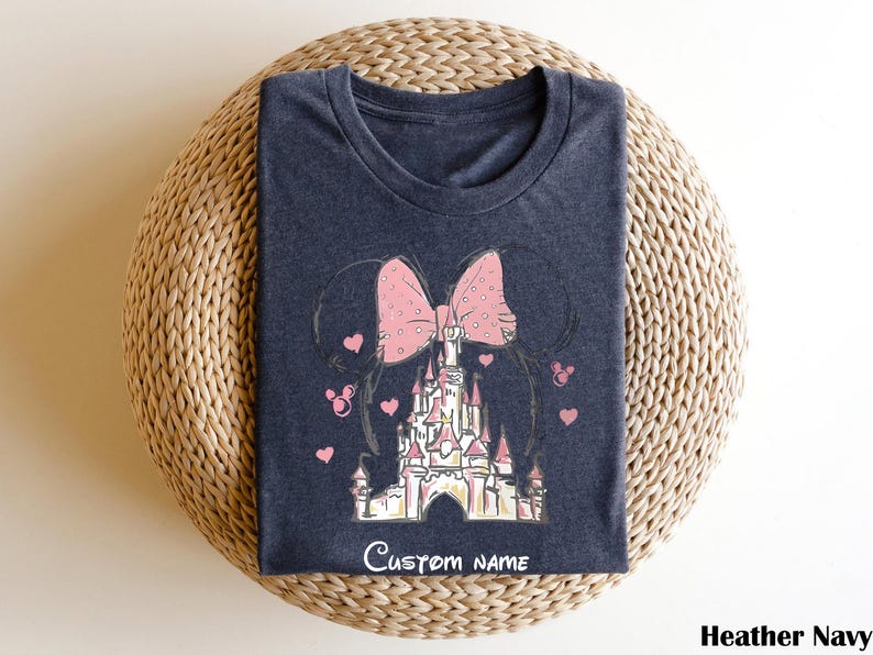 A dark blue t-shirt with a pink bow and a castle graphic printed on it, placed on a woven basket.