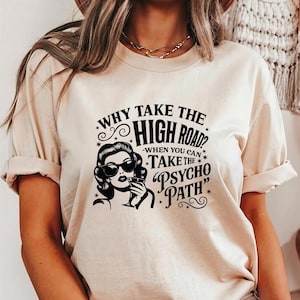May include: A beige t-shirt with rolled-up sleeves featuring a black graphic. The graphic includes text that reads "WHY TAKE THE HIGH ROAD? WHEN YOU CAN TAKE THE PSYCHO PATH" and an illustration of a woman wearing sunglasses.