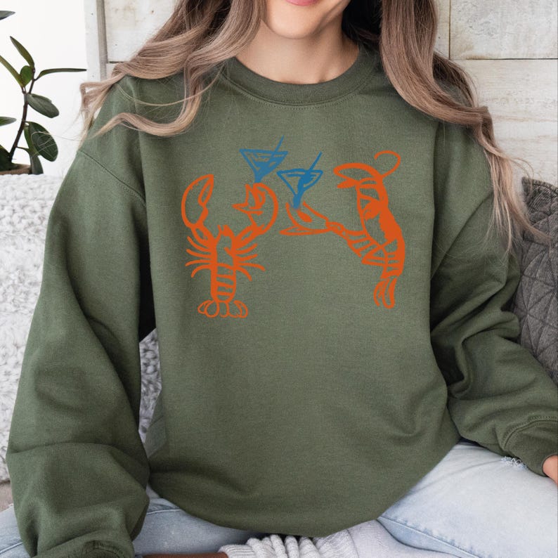May include: Olive green sweatshirt featuring an orange lobster graphic with two blue cocktail glasses. The graphic design is a fun, casual style. The sweatshirt is made of a soft material.