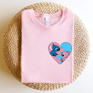 A pink t-shirt with a heart-shaped graphic featuring two cartoon characters, one blue and one pink, is displayed on a woven basket.