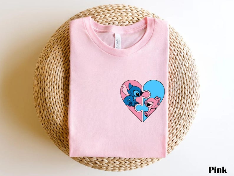 A pink t-shirt with a heart-shaped graphic featuring two cartoon characters, one blue and one pink, is displayed on a woven basket.