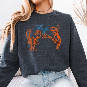 May include: Dark gray sweatshirt featuring an orange lobster design holding blue martini glasses. The graphic design is a playful illustration, perfect for casual wear. The sweatshirt is made of a soft material.