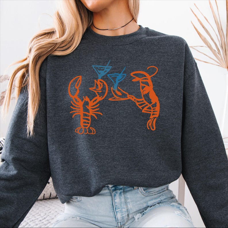 May include: Dark gray sweatshirt featuring an orange lobster design holding blue martini glasses. The graphic design is a playful illustration, perfect for casual wear. The sweatshirt is made of a soft material.