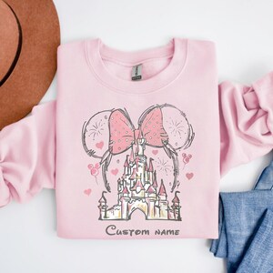 A pink sweatshirt with a castle and two pink bows on it, along with a brown hat and blue jeans, are laid out on a white surface.