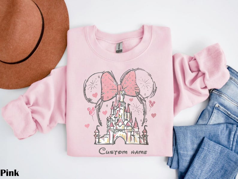 A pink sweatshirt with a castle and two pink bows on it, along with a brown hat and blue jeans, are laid out on a white surface.