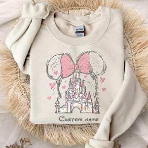 A beige sweater with a pink bow and a castle graphic printed on it, placed on a fluffy beige surface.