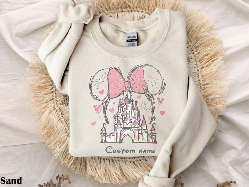 A beige sweater with a pink bow and a castle graphic printed on it, placed on a fluffy beige surface.