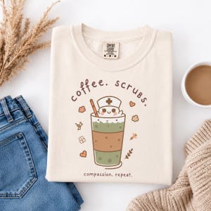 Op de afbeelding: Een crèmekleurig T-shirt met een afbeelding van een koffiedrankje met een verpleegsterskapje, omringd door kleine iconen zoals hartjes, een injectiespuit en pillen. De tekst coffee. scrubs. staat erboven en compassion. repeat. eronder.