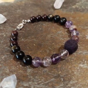 Peut inclure: Bracelet de perles avec un mélange de pierres précieuses rouge foncé, noires et violettes. Le bracelet comprend un groupe de perles violet foncé et un fermoir argenté. Les pierres précieuses varient en taille et en forme, créant un look unique et texturé.