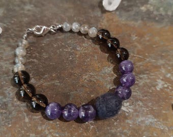 May include: A beaded bracelet featuring a variety of gemstones. The bracelet includes smoky quartz, amethyst, and other light-colored beads. A dark blue, porous stone is also included in the design. The bracelet is secured with a silver clasp.