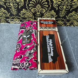 May include: A folded piece of bright pink fabric with a dark brown floral batik pattern, featuring a KOU brand tag, placed next to a rectangular wooden box with a lid labelled Authentic Indonesian Hand-Drawn Batik and Thank You For Your Order.