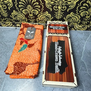 Può includere: Un tessuto arancione piegato con un motivo a foglie marroni e verde acqua, dotato di un'etichetta del marchio KOU, accanto a una scatola di legno rettangolare. Il coperchio riporta il testo Authentic Indonesian Hand-Drawn Batik e un'etichetta Thank You.