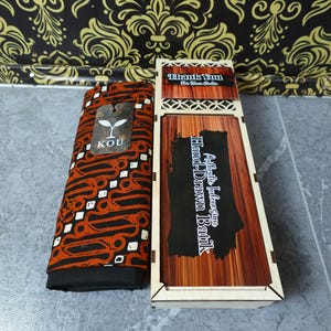 Puede incluir: Una pieza de tela doblada con un patrón batik marrón y negro y una etiqueta de marca KOU, junto a una caja de madera rectangular con tapa deslizante etiquetada como Authentic Indonesian Hand-Drawn Batik y una tarjeta de agradecimiento.