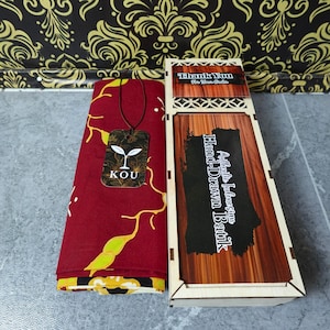 Puede incluir: Una pieza de tela roja doblada con un patrón floral amarillo y blanco, con una etiqueta que muestra el texto KOU y un icono de brote. Junto a ella hay una caja de madera rectangular con una tapa deslizante etiquetada Authentic Indonesian Hand-Drawn Batik.