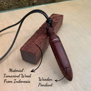 May include: A dark brown wooden pendant with a black cord, made from tamarind wood from Indonesia. The pendant is cylindrical with a silver band near the top. A rectangular piece of the same wood is behind the pendant.