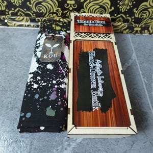 Könnte beinhalten: Ein gefalteter schwarzer Stoff mit weißen und violetten Farbspritzern und einem KOU-Markenanhänger neben einer rechteckigen Holzschachtel mit Holzmaserung. Der Deckel der Schachtel trägt den Text Authentic Indonesian Hand-Drawn Batik.