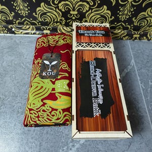 Può includere: Un tessuto rosso piegato con un motivo batik verde e giallo, dotato di un'etichetta del marchio KOU, accanto a una scatola di legno rettangolare con coperchio scorrevole. La scatola riporta il testo Thank You For Your Order e Authentic Indonesian Hand-Drawn Batik.