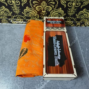 Könnte beinhalten: Ein gerollter orangefarbener Stoff mit Blatt- und Fruchtmuster neben einer rechteckigen Holzschachtel. Der Deckel der Schachtel zeigt den Text "Authentic Indonesian Hand-Drawn Batik" und ein "Thank You For Your Order" Etikett.