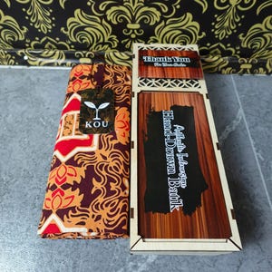 Könnte beinhalten: Ein gefaltetes Stoffstück mit einem batikartigen Muster in Rot, Orange und Dunkelbraun mit einem KOU-Markenetikett, neben einer rechteckigen Holzschachtel mit Schiebedeckel, beschriftet mit Authentic Indonesian Hand-Drawn Batik und Thank You For Your Order.